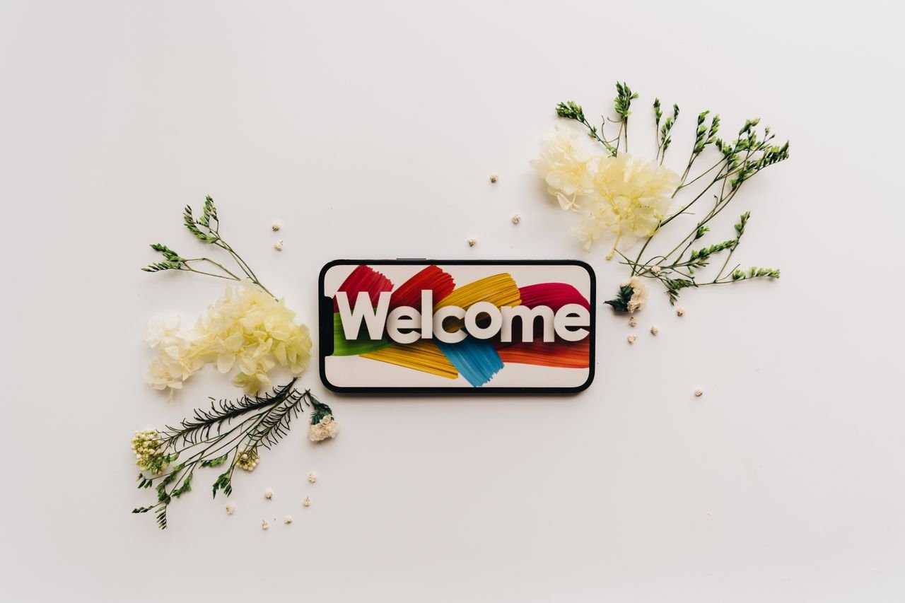 Creative flat lay with a smartphone displaying a colorful welcome sign surrounded by delicate flowers.